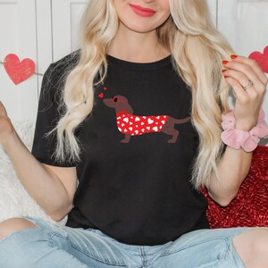 May include: A black t-shirt with a red dachshund wearing a red heart-patterned sweater and sunglasses. The dachshund is blowing hearts.