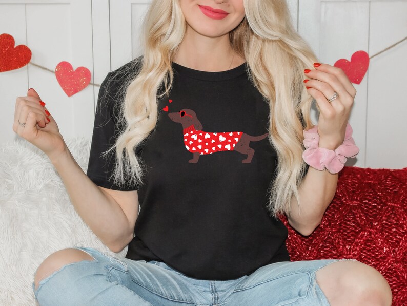 May include: A black t-shirt with a red dachshund wearing a red heart-patterned sweater and sunglasses. The dachshund is blowing hearts.