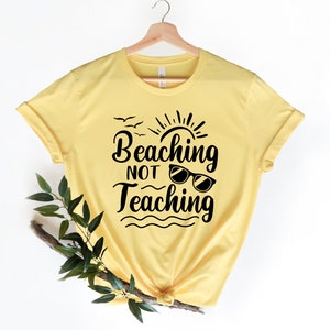 Op de afbeelding: Geel T-shirt met een zwarte afbeelding waarop staat "Beaching Not Teaching" met een zon, vogels en een zonnebril.