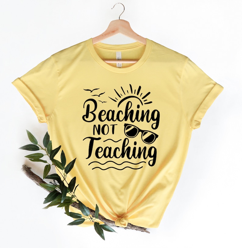 Op de afbeelding: Geel T-shirt met een zwarte afbeelding waarop staat "Beaching Not Teaching" met een zon, vogels en een zonnebril.