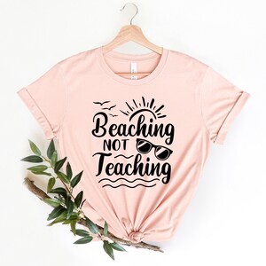 Op de afbeelding: Een roze T-shirt met de tekst "Beaching Not Teaching" in zwarte letters. De tekst is gestyled met een zon, vogels en een zonnebril.