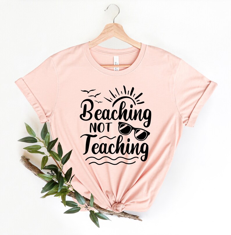 Op de afbeelding: Een roze T-shirt met de tekst "Beaching Not Teaching" in zwarte letters. De tekst is gestyled met een zon, vogels en een zonnebril.