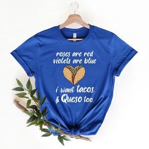 Op de afbeelding: Een koninklijk blauw t-shirt met een witte afbeelding waarop staat "roses are red violets are blue i want tacos & queso too." De afbeelding bevat ook een hartvorm gemaakt van twee taco's.