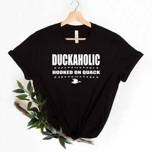 May include: Black t-shirt with white text that reads "Duckaholic Hooked on Quack" and a white silhouette of a duck.