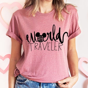 May include: A pink t-shirt with a black graphic that says "world traveler" with a Mickey Mouse head and a globe inside the ears.