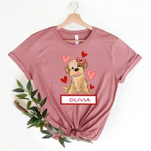 May include: A mauve t-shirt with a cartoon image of a brown dog wearing a red bow with white polka dots. The dog is surrounded by red hearts. The text "OLIVIA" is printed on a red banner below the dog.
