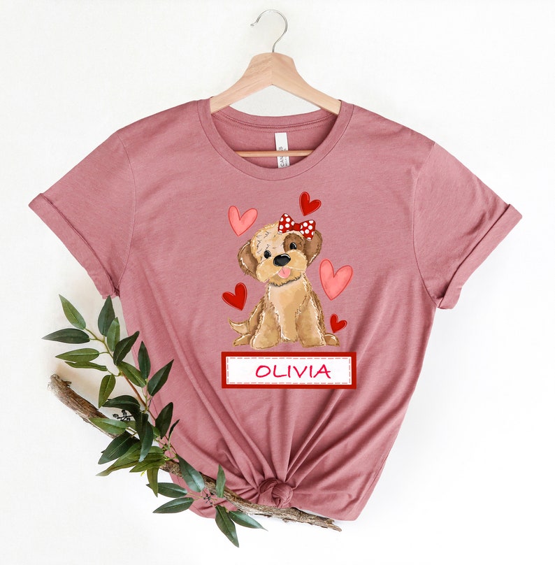 May include: A mauve t-shirt with a cartoon image of a brown dog wearing a red bow with white polka dots. The dog is surrounded by red hearts. The text "OLIVIA" is printed on a red banner below the dog.