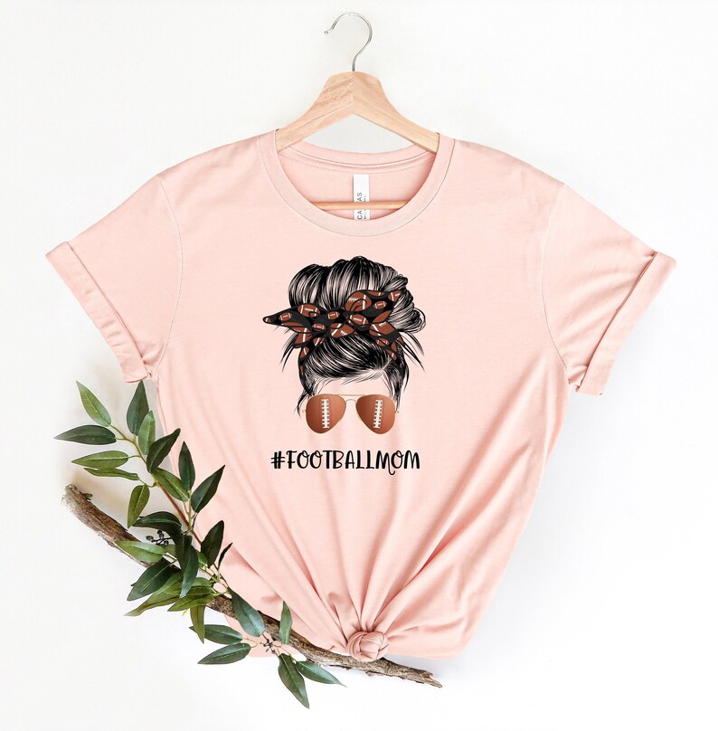 May include: A pink t-shirt with a graphic of a woman with a messy bun, wearing sunglasses with footballs on the lenses. The bun is decorated with a bandana with footballs. The text "#FOOTBALLMOM" is printed below the graphic.