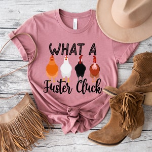K&ouml;nnte beinhalten: Ein pinkfarbenes T-Shirt mit dem Text "What a Fuster Cluck" und einer Reihe von f&uuml;nf H&uuml;hnern, die auf der Vorderseite aufgedruckt sind. Das Shirt ist unten verknotet.