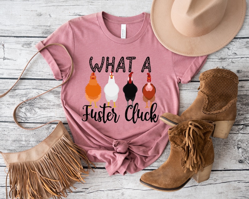 K&ouml;nnte beinhalten: Ein pinkfarbenes T-Shirt mit dem Text "What a Fuster Cluck" und einer Reihe von f&uuml;nf H&uuml;hnern, die auf der Vorderseite aufgedruckt sind. Das Shirt ist unten verknotet.