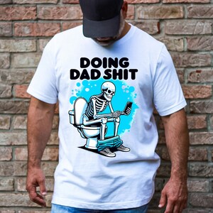 May include: White t-shirt with a skeleton sitting on a toilet and holding a phone. The text "Doing Dad Shit" is printed on the shirt.
