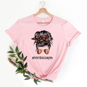 May include: Pink t-shirt with a graphic of a woman with a messy bun, wearing sunglasses and a headband with footballs. The text "#FOOTBALLMOM" is printed below the graphic.