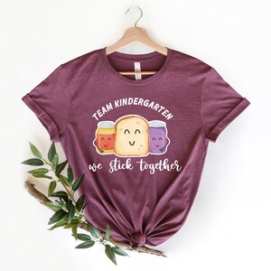 May include: A maroon t-shirt with the text "Team Kindergarten we stick together" and a graphic of three cartoon jars of peanut butter, jelly, and jam with a slice of bread.