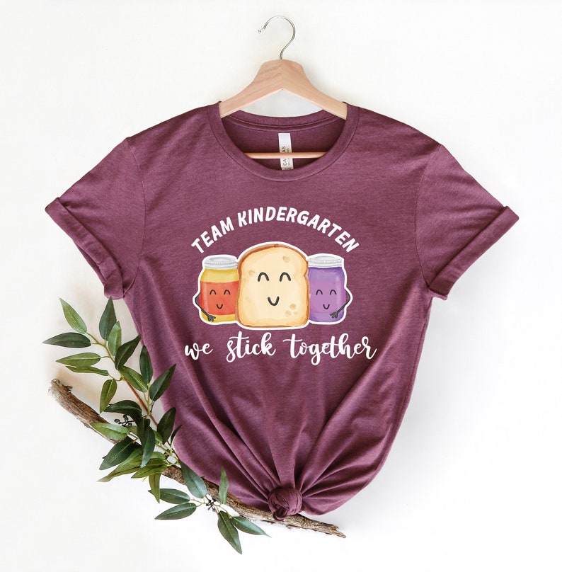 May include: A maroon t-shirt with the text "Team Kindergarten we stick together" and a graphic of three cartoon jars of peanut butter, jelly, and jam with a slice of bread.