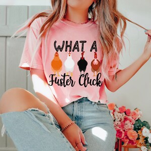 K&ouml;nnte beinhalten: Ein pinkfarbenes T-Shirt mit dem Text "What a Fuster Cluck" und einer Reihe von f&uuml;nf H&uuml;hnern, die auf der Vorderseite aufgedruckt sind.
