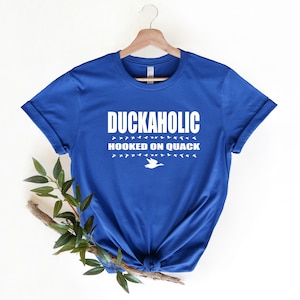 May include: A royal blue t-shirt with white text that reads "Duckaholic Hooked on Quack" and a white silhouette of a duck flying above the text.