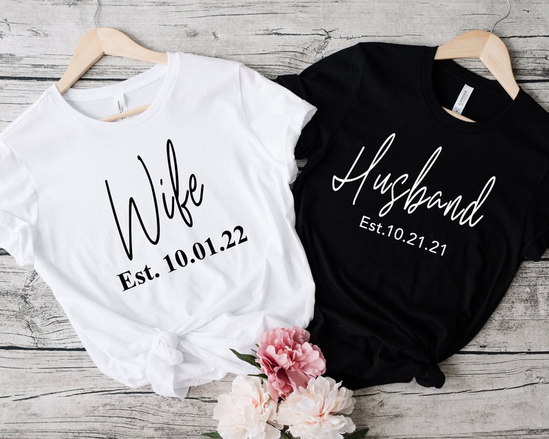 May include: Two white t-shirts with black text. One shirt says "Wife Est. 10.01.22" and the other says "Husband Est. 10.21.21".
