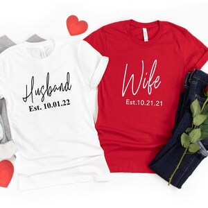 May include: Two white t-shirts with black text. One says "Husband Est. 10.01.22" and the other says "Wife Est. 10.21.21".