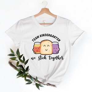 May include: White t-shirt with a black graphic of a slice of bread with two jars of jam on either side. The text "Team Kindergarten we stick together" is printed below the graphic.