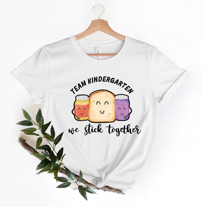 May include: White t-shirt with a black graphic of a slice of bread with two jars of jam on either side. The text "Team Kindergarten we stick together" is printed below the graphic.
