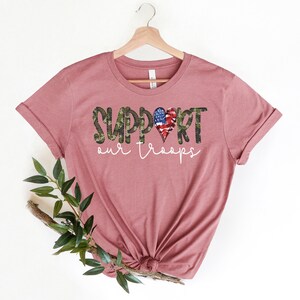 May include: A mauve t-shirt with a graphic design of the word "SUPPORT" in camouflage print with an American flag heart in the middle of the word. The text "our troops" is printed below the word "SUPPORT".