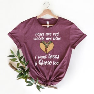 Op de afbeelding: Bordeauxkleurig T-shirt met witte tekst die "Roses are red, violets are blue, I want tacos & queso too." luidt. De tekst wordt vergezeld door een afbeelding van twee taco's in de vorm van een hart.