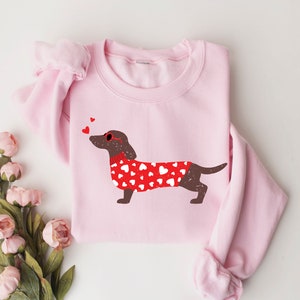 May include: Pink sweatshirt with a brown dachshund wearing a red sweater with white hearts and red heart-shaped sunglasses. Three small red hearts are above the dachshund.