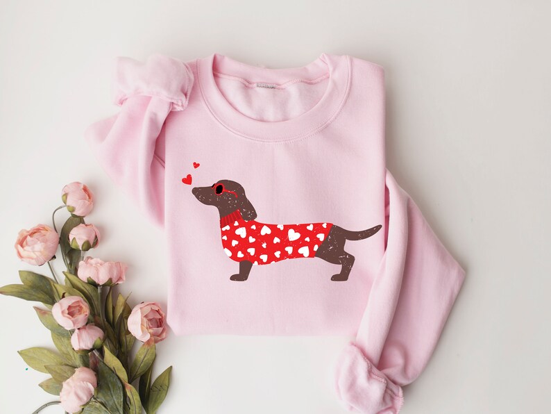 May include: Pink sweatshirt with a brown dachshund wearing a red sweater with white hearts and red heart-shaped sunglasses. Three small red hearts are above the dachshund.