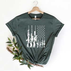 May include: A dark green t-shirt with a white distressed American flag design featuring a row of guns.