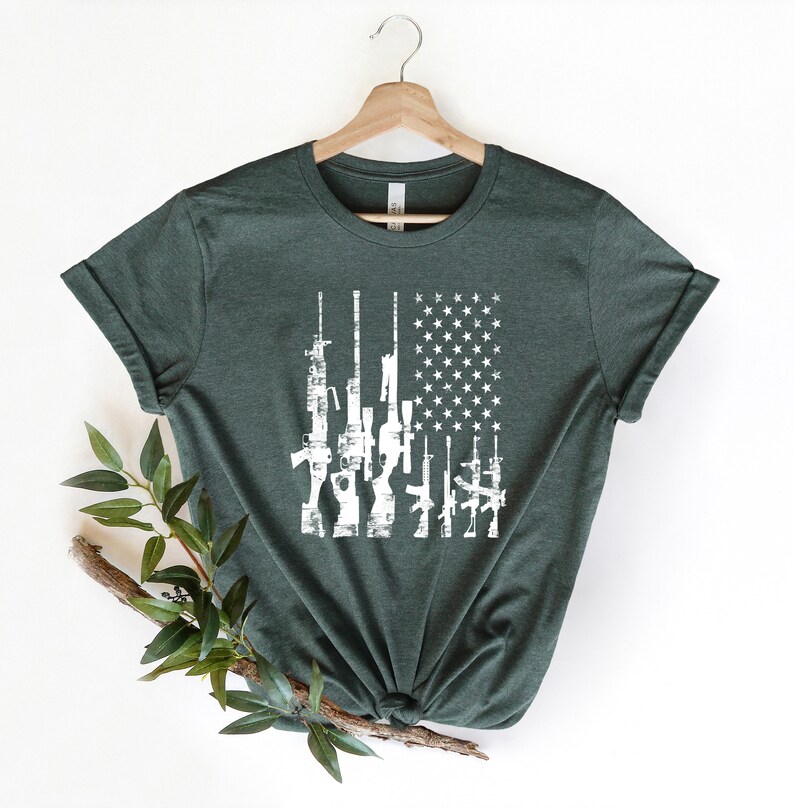 May include: A dark green t-shirt with a white distressed American flag design featuring a row of guns.