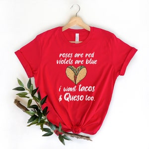 Op de afbeelding: Rood T-shirt met een witte afbeelding waarop staat "roses are red violets are blue i want tacos & queso too." De afbeelding bevat ook een hartvormig beeld van twee taco's.
