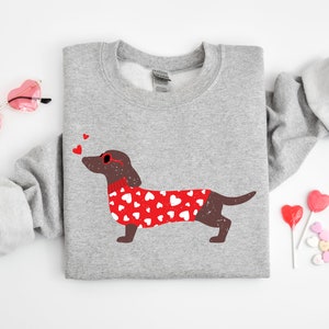 May include: A gray sweatshirt with a brown dachshund wearing red heart-patterned sweater and red heart-shaped sunglasses. The dachshund is blowing a kiss. There are three red hearts above the dachshund's head.