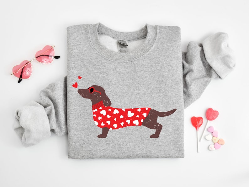 May include: A gray sweatshirt with a brown dachshund wearing red heart-patterned sweater and red heart-shaped sunglasses. The dachshund is blowing a kiss. There are three red hearts above the dachshund's head.