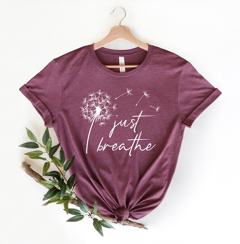 Pu&ograve; includere: Una maglietta color borgogna con un motivo di dente di leone bianco e il testo "just breathe".
