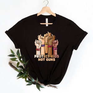 May include: Black t-shirt with a graphic of five raised fists in different skin tones, each wearing colorful bracelets. The text "PROTECT KIDS NOT GUNS" is printed below the fists.