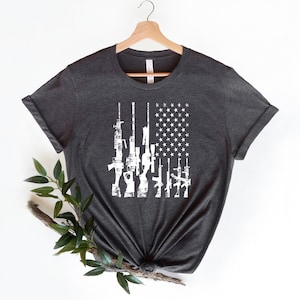 May include: A dark heather grey t-shirt with a white distressed design of an American flag with guns in place of the stripes.