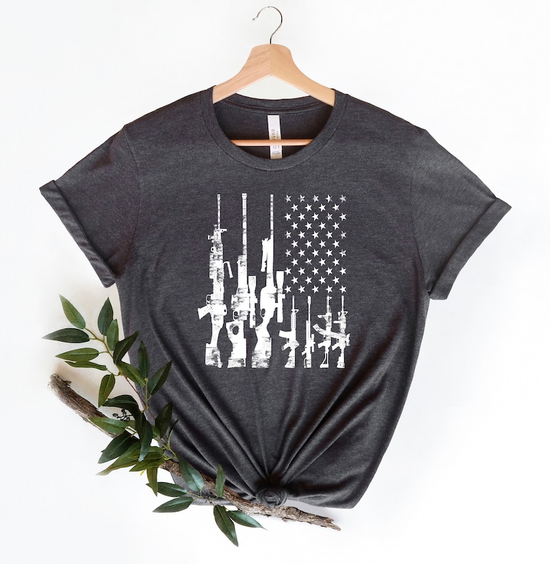 May include: A dark heather grey t-shirt with a white distressed design of an American flag with guns in place of the stripes.