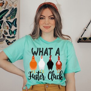 K&ouml;nnte beinhalten: Ein t&uuml;rkisfarbenes T-Shirt mit dem Aufdruck "WHAT A Fuster Cluck" auf der Vorderseite. Der Text ist schwarz mit wei&szlig;em Umriss. Unter dem Text befinden sich vier Cartoon-H&uuml;hner in einer Reihe. Die H&uuml;hner sind orange, wei&szlig;, schwarz und braun.