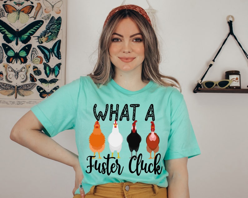 K&ouml;nnte beinhalten: Ein t&uuml;rkisfarbenes T-Shirt mit dem Aufdruck "WHAT A Fuster Cluck" auf der Vorderseite. Der Text ist schwarz mit wei&szlig;em Umriss. Unter dem Text befinden sich vier Cartoon-H&uuml;hner in einer Reihe. Die H&uuml;hner sind orange, wei&szlig;, schwarz und braun.
