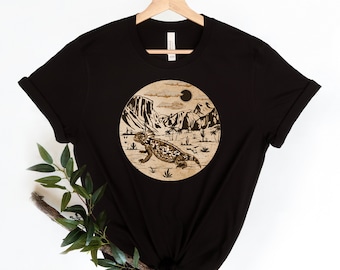May include: Black t-shirt with a circular graphic of a desert scene with a lizard, mountains, and a sun.