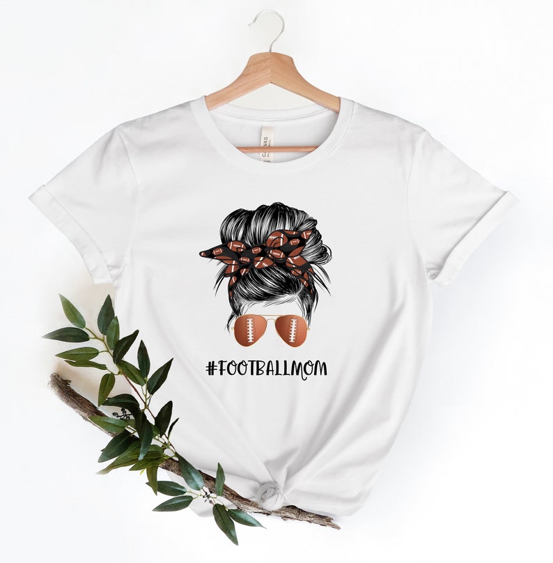 May include: White t-shirt with a graphic of a woman with a messy bun, wearing sunglasses with footballs on the lenses. The bun is decorated with a bandana with footballs. The text "#FOOTBALLMOM" is printed below the graphic.