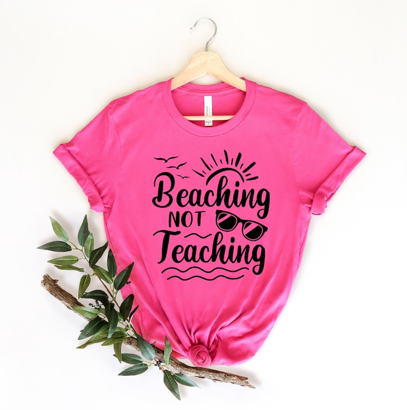 Op de afbeelding: Een roze T-shirt met een zwarte afbeelding waarop staat "Beaching Not Teaching" met een zon, vogels en een zonnebril.