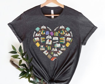 Può includere: T-shirt grigio scuro con un design a forma di cuore composto da vari libri, tazze da tè ed elementi floreali. Il design include libri aperti e chiusi in vari colori, creando un tema letterario. La maglietta è appesa a una gruccia di legno.