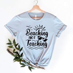 Op de afbeelding: Lichtblauw T-shirt met een zwarte afbeelding met de tekst "Beaching Not Teaching" met een zon, vogels en een zonnebril.