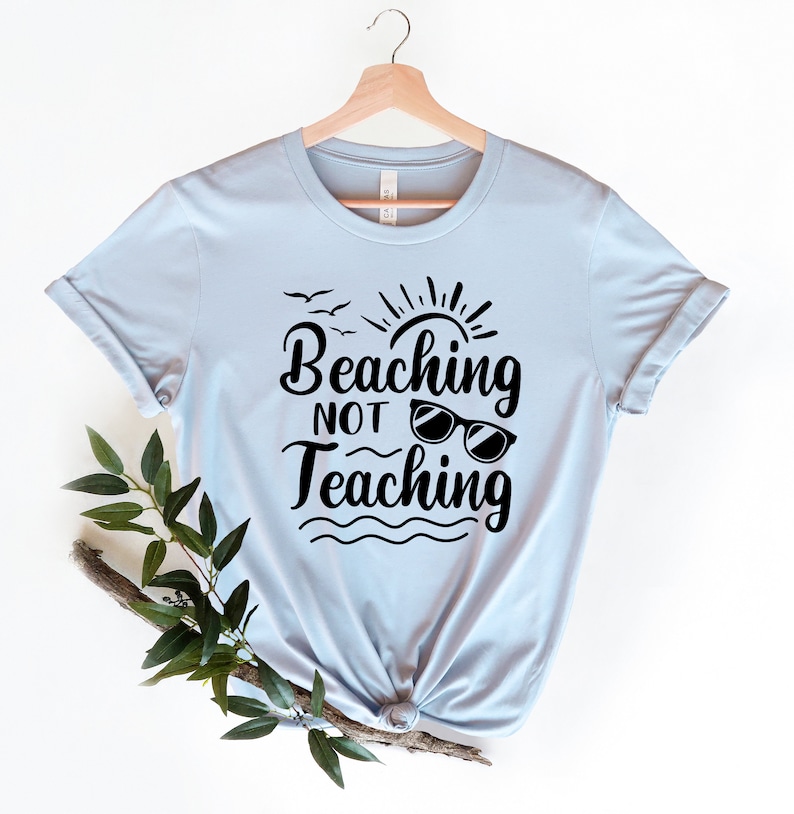 Op de afbeelding: Lichtblauw T-shirt met een zwarte afbeelding met de tekst "Beaching Not Teaching" met een zon, vogels en een zonnebril.