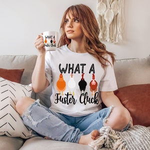 K&ouml;nnte beinhalten: Ein wei&szlig;es T-Shirt mit dem Aufdruck "What a Fuster Cluck". Der Text ist von einer Reihe schwarzer Punkte umgeben. Unter dem Text befinden sich f&uuml;nf Cartoon-H&uuml;hner in einer Reihe. Die H&uuml;hner sind orange, wei&szlig;, schwarz, braun und orange. Die Person, die das Shirt tr&auml;gt, sitzt auf einem Sofa.