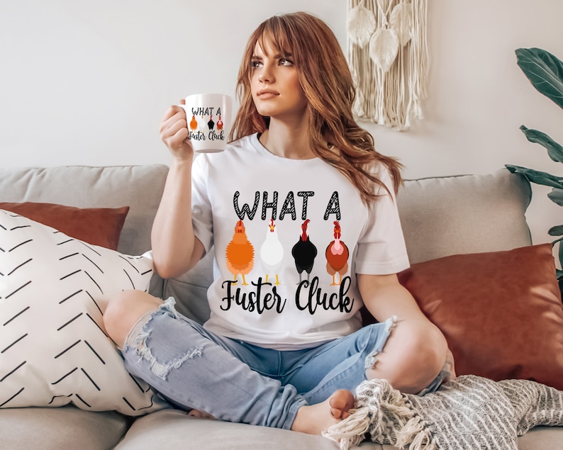 K&ouml;nnte beinhalten: Ein wei&szlig;es T-Shirt mit dem Aufdruck "What a Fuster Cluck". Der Text ist von einer Reihe schwarzer Punkte umgeben. Unter dem Text befinden sich f&uuml;nf Cartoon-H&uuml;hner in einer Reihe. Die H&uuml;hner sind orange, wei&szlig;, schwarz, braun und orange. Die Person, die das Shirt tr&auml;gt, sitzt auf einem Sofa.