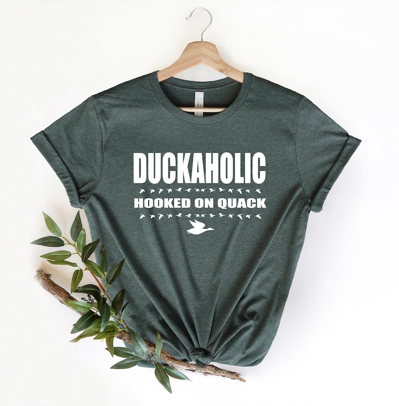 May include: A dark green t-shirt with the text "Duckaholic Hooked on Quack" in white. The text is surrounded by a row of white duck silhouettes.