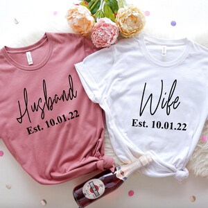 May include: Two white t-shirts with black text. The left shirt is a dusty rose color and says "Husband Est. 10.01.22". The right shirt is white and says "Wife Est. 10.01.22".