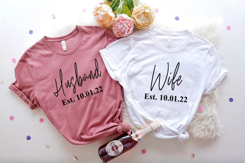 May include: Two white t-shirts with black text. The left shirt is a dusty rose color and says "Husband Est. 10.01.22". The right shirt is white and says "Wife Est. 10.01.22".